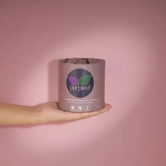 Hand holding a roll of Purpleef biodegradable toilet paper against a pink background